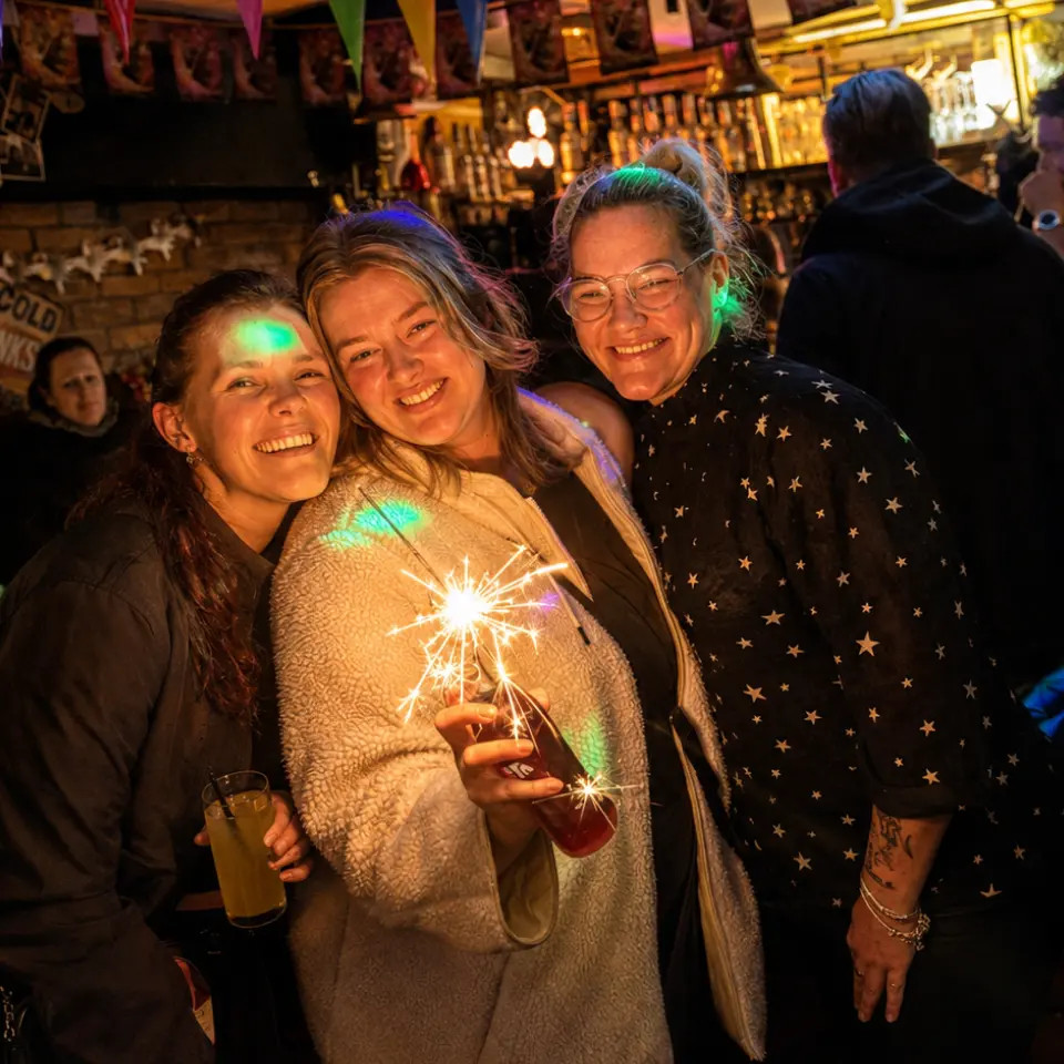 Drie vrienden poseren in een bar met kleurrijke verlichting, lachend en met vonkende drankjes.